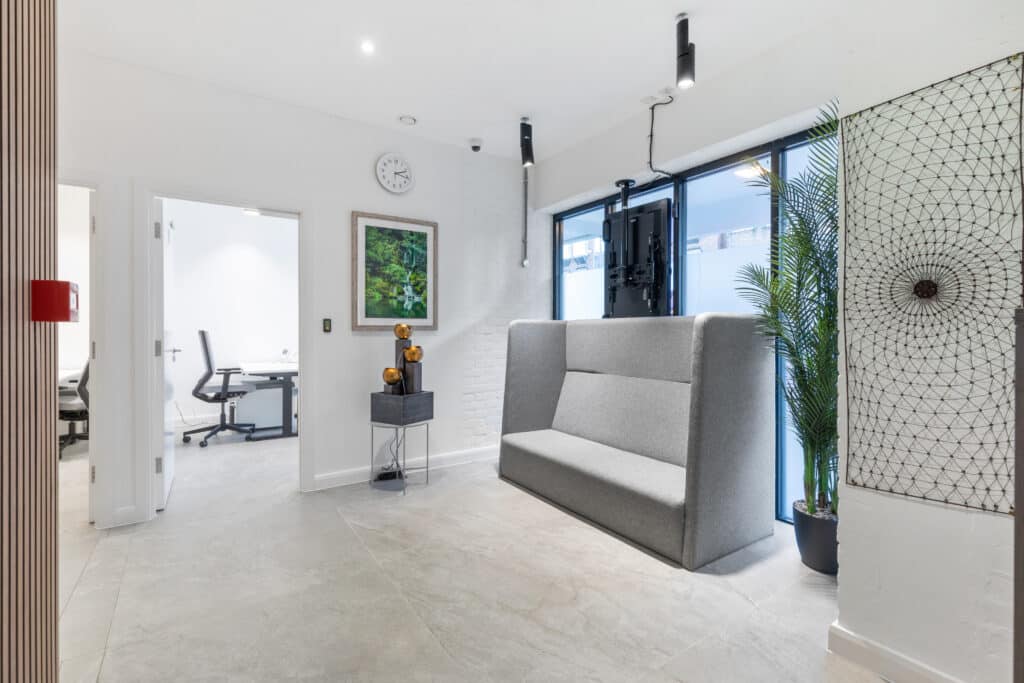 The modern office entrance features a grey cushioned bench against a white brick wall, embodying the "About Us" essence. A tall potted plant adds vibrancy next to the bench. Abstract art adorns the space alongside a side table with decor. Natural light floods through large windows, brightening the scene.