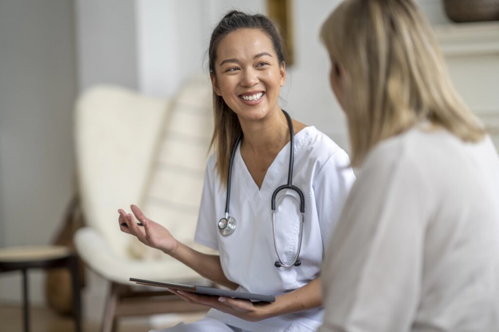 In our "About Us" section, a healthcare professional in white scrubs, stethoscope around her neck, warmly converses with a patient. Holding a tablet in the softly lit consultation area, she ensures each patient's comfort and trust.