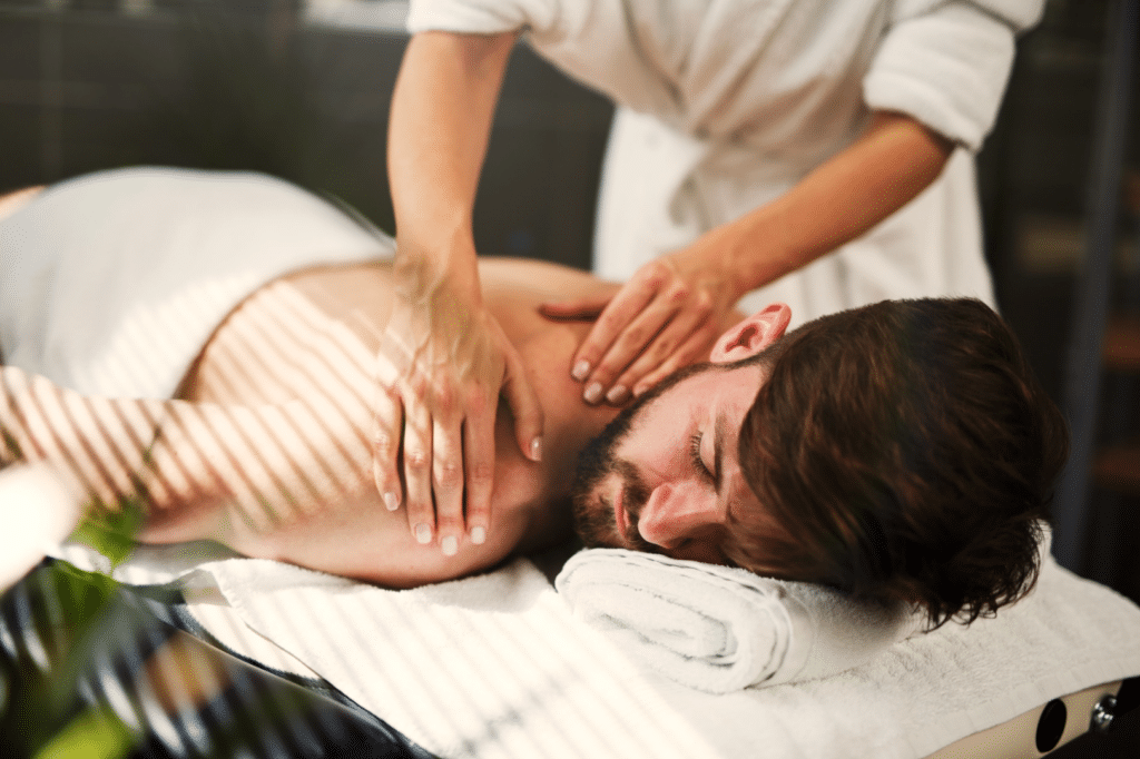 A man with a beard lies face down on a massage table, eyes closed, wrapped in a white towel. An individual in a robe massages his shoulders with expert hands. Sunlight filters through blinds, casting striped shadows on the scene, creating a serene atmosphere that speaks volumes about us.