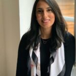 A person with long dark hair is sitting by a window, smiling. They are wearing a black top and a white scarf with black and pink accents. The background reveals a blurred view of greenery outside the window, embodying the serenity of health psychology akin to Dr. Puja Patel's approach.