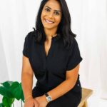 A woman with shoulder-length black hair and a black blouse smiles while sitting on a wooden stool. Chanel Tamana wears black pants and a watch on her left wrist. Green leaves are beside her, set against a white curtain background with a woven rug on the floor.