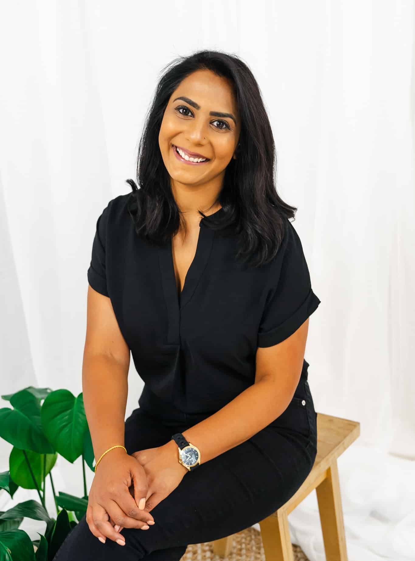 A woman with shoulder-length black hair and a black blouse smiles while sitting on a wooden stool. Chanel Tamana wears black pants and a watch on her left wrist. Green leaves are beside her, set against a white curtain background with a woven rug on the floor.