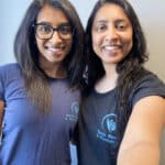 Two women with long dark hair stand close together, smiling at the camera. Both wear navy or black T-shirts with a blue Nanda Physio logo and “Nanda Massage & Physiotherapy” text, highlighting their dedication to physiotherapy. The background is plain and light-colored.