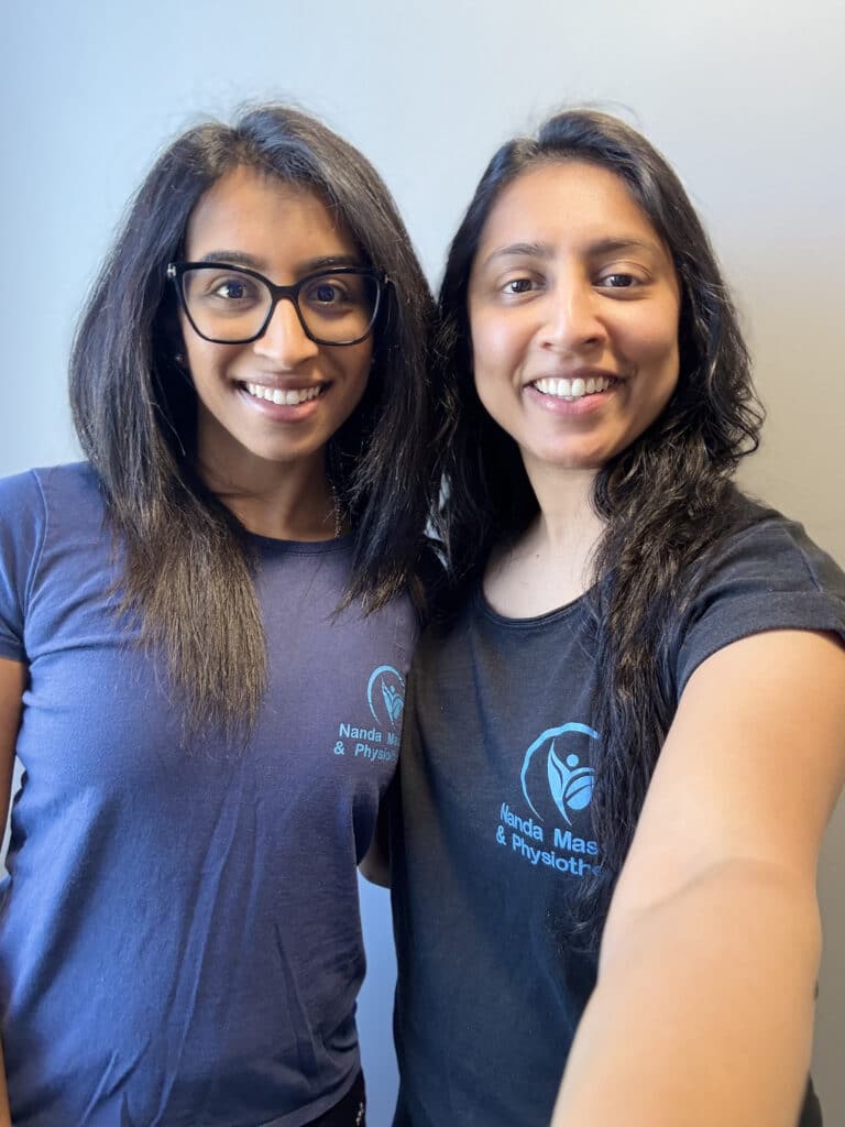 Two women with long dark hair stand close together, smiling at the camera. Both wear navy or black T-shirts with a blue Nanda Physio logo and “Nanda Massage & Physiotherapy” text, highlighting their dedication to physiotherapy. The background is plain and light-colored.