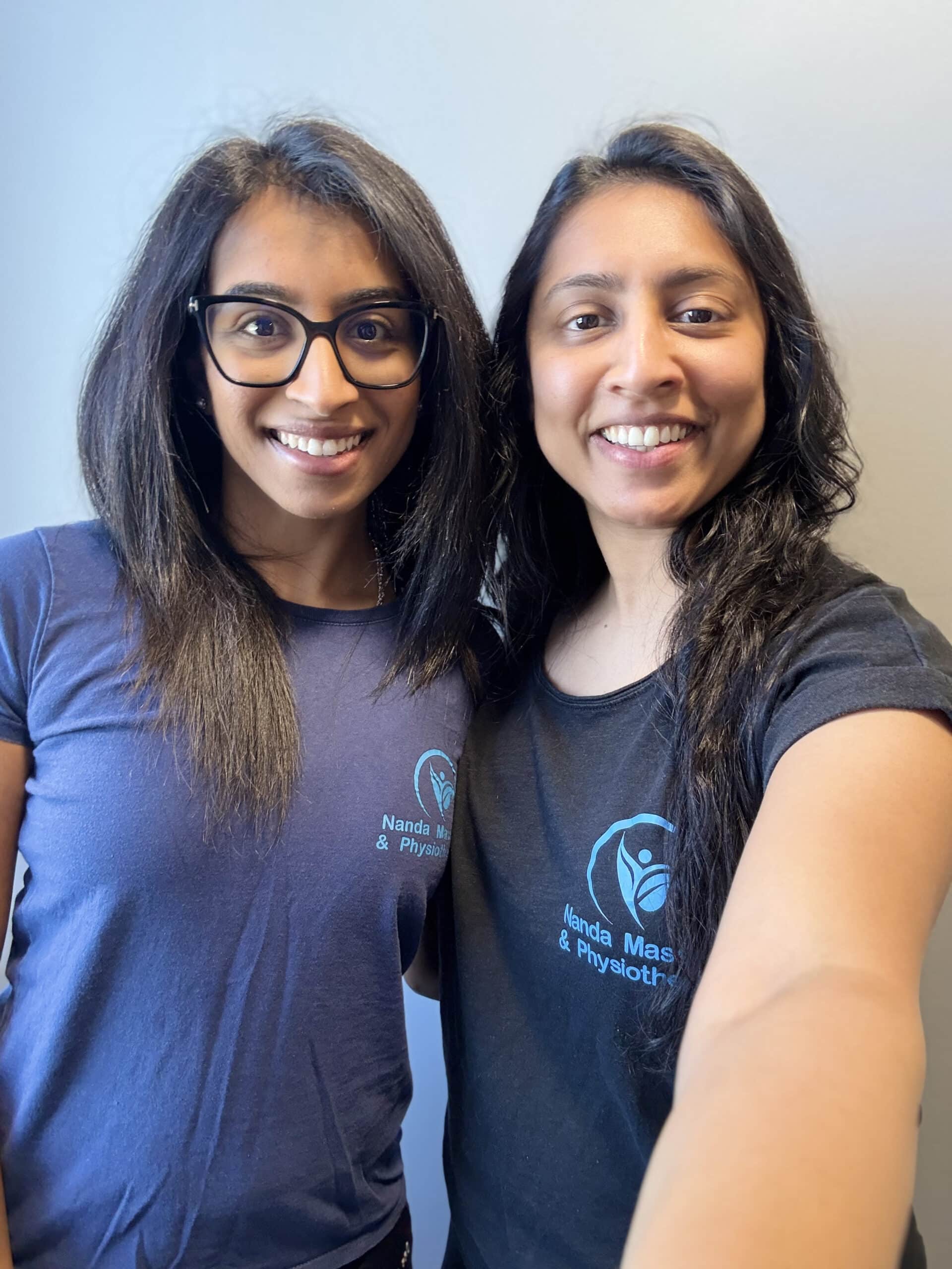 Two women with long dark hair stand close together, smiling at the camera. Both wear navy or black T-shirts with a blue Nanda Physio logo and “Nanda Massage & Physiotherapy” text, highlighting their dedication to physiotherapy. The background is plain and light-colored.