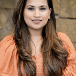 A woman with long, wavy brown hair smiles at the camera. She is wearing a peach, textured blouse with puffed sleeves. Her jewelry includes small earrings and a pendant necklace. The background is a stone wall, slightly out of focus, which provides a neutral backdrop.