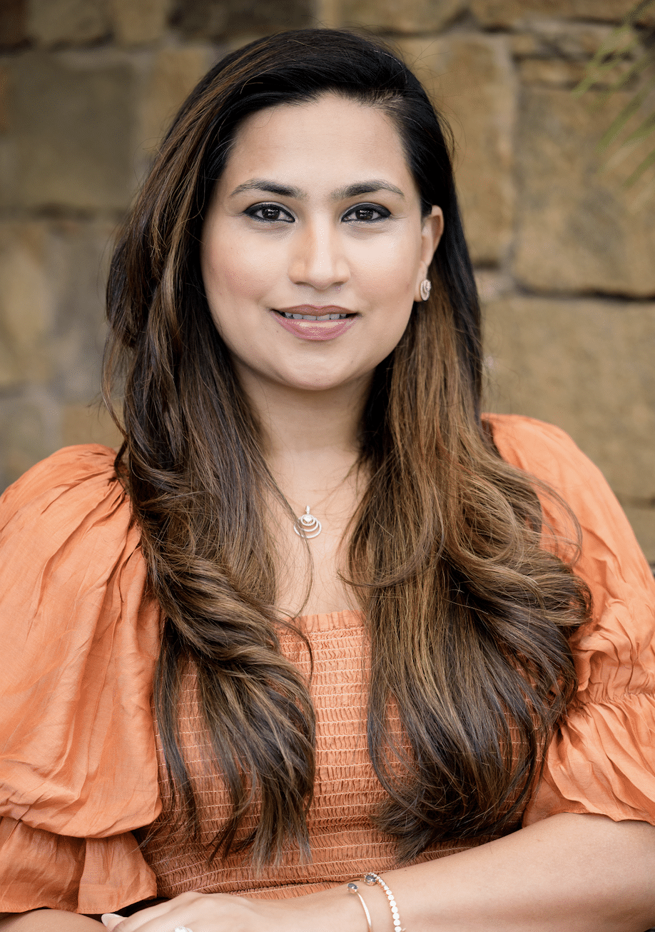 A woman with long, wavy brown hair smiles at the camera. She is wearing a peach, textured blouse with puffed sleeves. Her jewelry includes small earrings and a pendant necklace. The background is a stone wall, slightly out of focus, which provides a neutral backdrop.