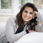 Sabrina Khaliya, with her long, wavy brown hair, smiles as she leans on a white sofa. She's wearing a gray cardigan over a black top. The blurred windows and greenery in the background create a bright and relaxed atmosphere.