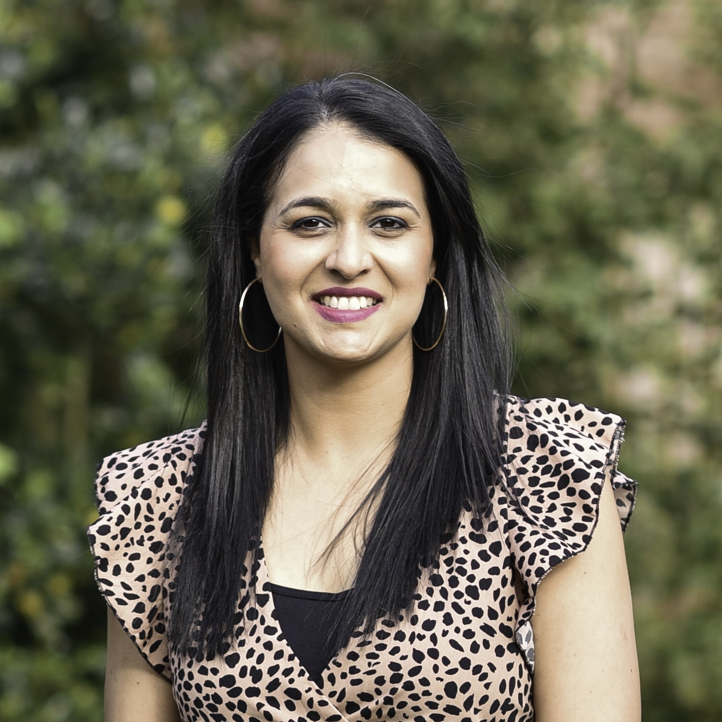 A smiling woman with long black hair stands outdoors. She's wearing a pink blouse with black polka dots and hoop earrings. The background features green foliage, slightly blurred, suggesting a natural setting. Meet Maya Mistry, your friendly neighborhood podiatrist enjoying a moment in nature's embrace.