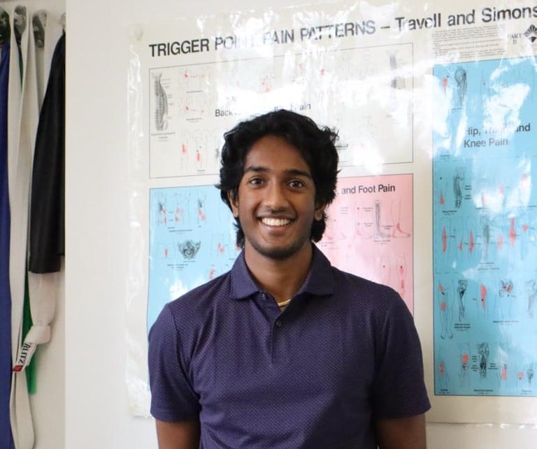 A person with dark, curly hair stands smiling in front of a wall adorned with anatomical charts, highlighting diagrams related to trigger point pain patterns. Several belts hang on the left side. Sporting a dark purple polo shirt, they embody the professionalism of Performax Chiropractic care.