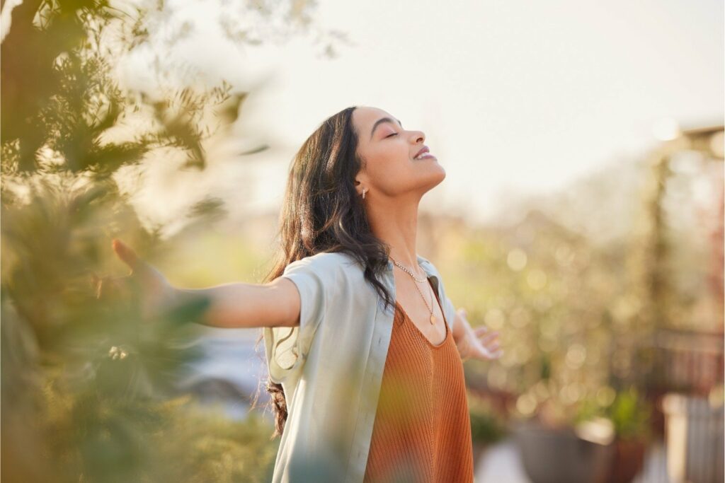 A woman stands outdoors with her arms outstretched and eyes closed, smiling peacefully. She wears a light blue shirt over an orange top. Sunlight softly illuminates her face as she enjoys the greenery, radiating joy and physical well-being.