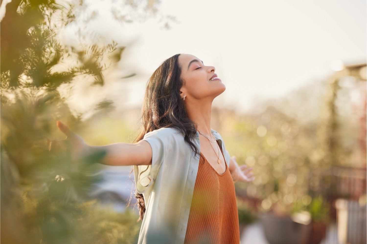 A woman stands outdoors with her arms outstretched and eyes closed, smiling peacefully. She wears a light blue shirt over an orange top. Sunlight softly illuminates her face as she enjoys the greenery, radiating joy and physical well-being.