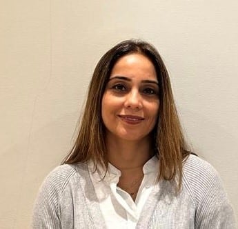A woman with straight, shoulder-length brown hair is smiling softly. She wears a light gray sweater over a white collared shirt, standing against a beige wall. Her warm, approachable mood lets her Words Express kindness and confidence.