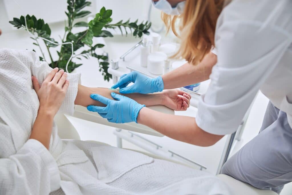 A healthcare worker wearing blue gloves prepares to administer a Myers’ Cocktail IV Drip Therapy to a patient’s arm. The patient is lying on a white medical chair in a white robe, with medical supplies and a green plant visible in the background.