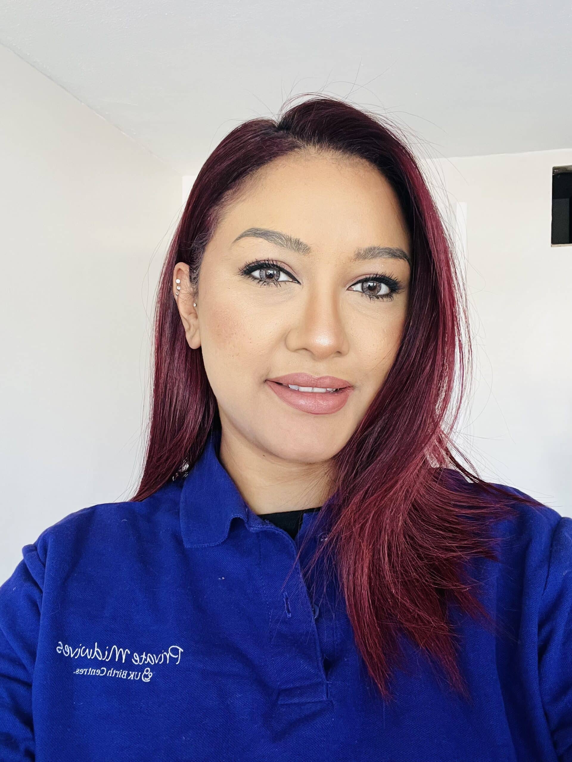 A woman with straight, shoulder-length dark red hair smiles at the camera. She is wearing a royal blue collared shirt with white text for Private Midwives. The background is a brightly lit, plain white room.