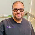 A man with a beard and glasses smiles while wearing black scrubs embroidered with “The Pharmaesthetics Clinic.” He stands indoors in front of a modern staircase at this leading aesthetics clinic, framed by off-white walls and vertical lights.