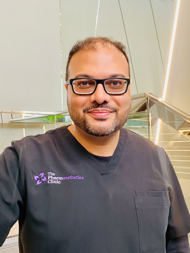 A man with a beard and glasses smiles while wearing black scrubs embroidered with “The Pharmaesthetics Clinic.” He stands indoors in front of a modern staircase at this leading aesthetics clinic, framed by off-white walls and vertical lights.