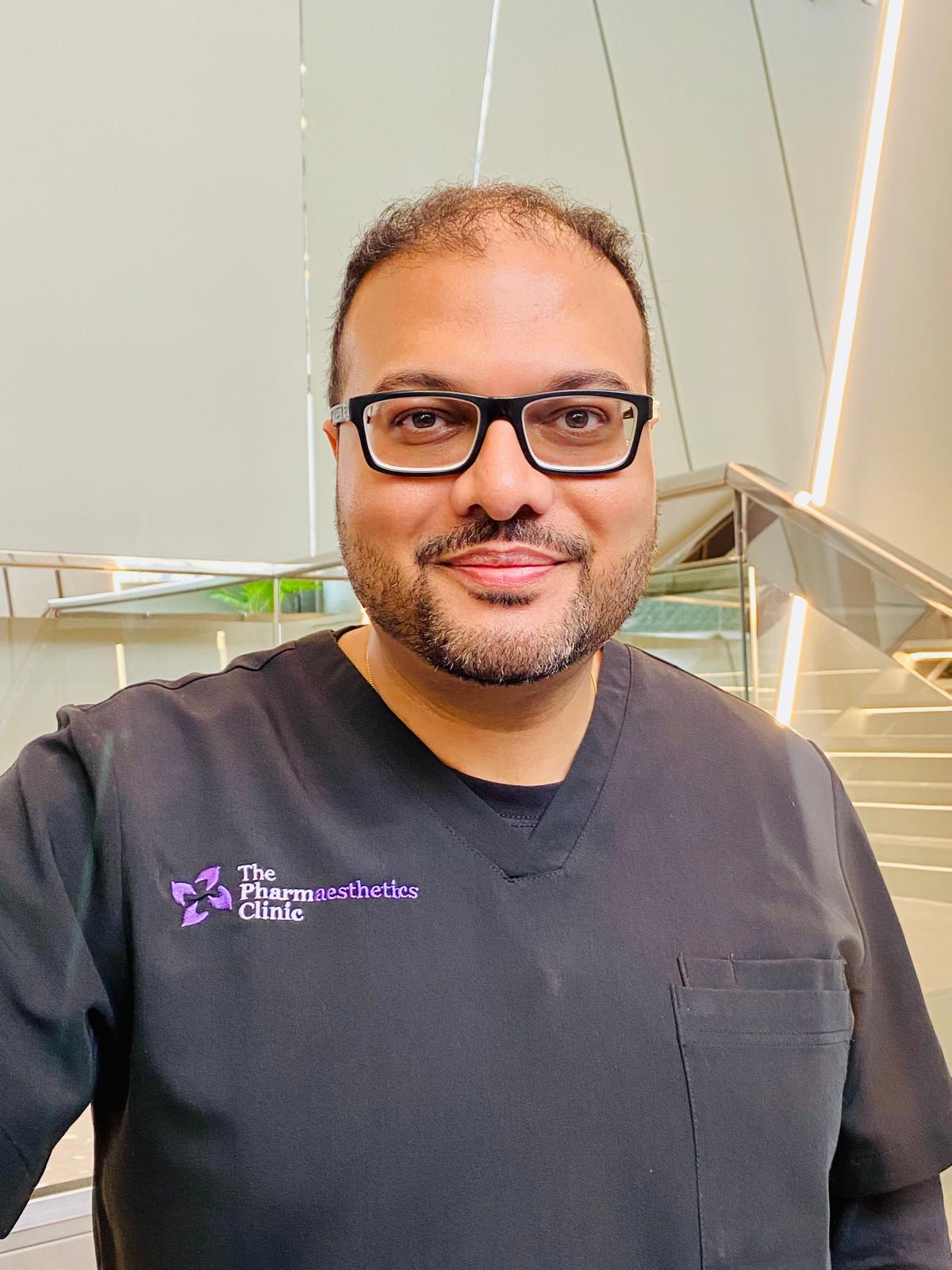 A man with a beard and glasses smiles while wearing black scrubs embroidered with “The Pharmaesthetics Clinic.” He stands indoors in front of a modern staircase at this leading aesthetics clinic, framed by off-white walls and vertical lights.