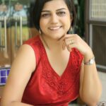 Nidhi Samani, a woman with short dark hair, smiles while sitting at a patterned table in a sleeveless red dress with floral embroidery. She rests her chin on her hand, wearing a watch and gold ring, with glass doors and greenery in the background.