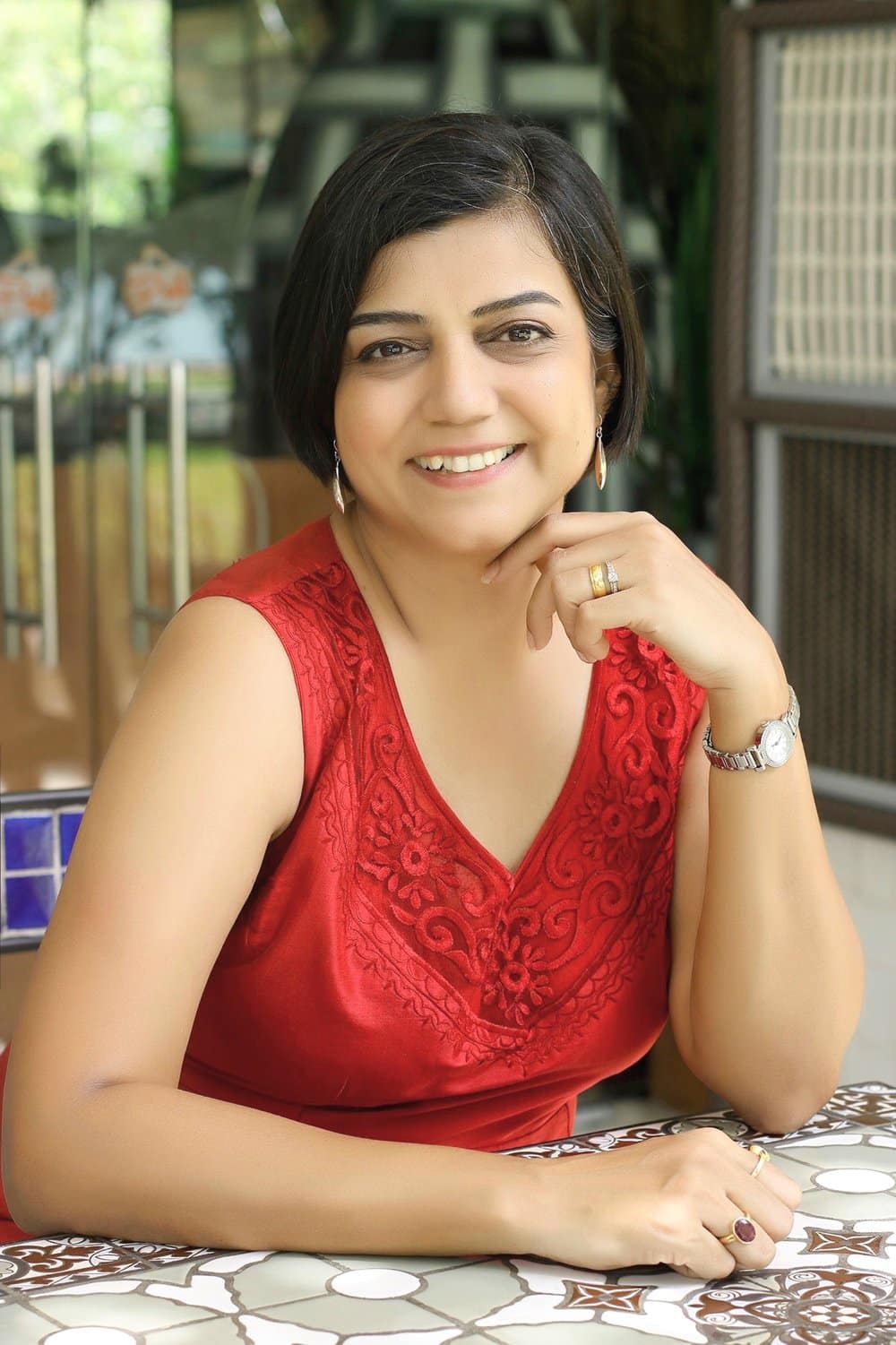 Nidhi Samani, a woman with short dark hair, smiles while sitting at a patterned table in a sleeveless red dress with floral embroidery. She rests her chin on her hand, wearing a watch and gold ring, with glass doors and greenery in the background.